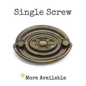 May include: An antique brass drawer pull with an oval shape and a floral design in the center. The words "Single Screw" are above the pull, and "More Available" is below it.
