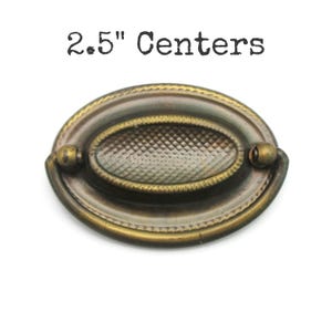 May include: Oval-shaped antique brass drawer pull with a textured center and decorative trim. The pull has a 2.5-inch center-to-center measurement and is set against a white background.