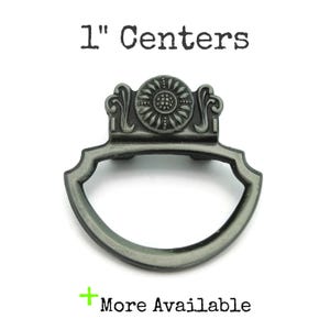 May include: A gray metal drawer pull with a decorative floral design and a curved handle. The text "1" Centers" is above the pull, and "More Available" is below. The pull measures 1 inch across the center.