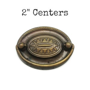 May include: Antique brass drawer pull with a decorative oval center and a handle. The pull has a 2-inch center-to-center measurement. The oval center features a leaf design. The pull is made of metal and has a vintage look.