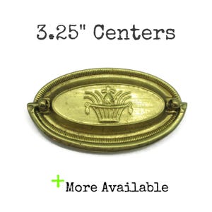 May include: A brass-toned oval drawer pull with a decorative basket design in the center. The pull has a ridged border and small circular accents on each end. The text "3.25" Centers" is above the pull.