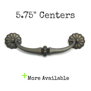 May include: A dark bronze-toned metal drawer pull with a curved handle and two circular, flower-shaped mounting plates. The text 5.75" Centers is printed above the handle, and the text + More Available is printed below the handle.