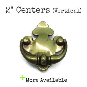 May include: A brass-toned drawer pull with a decorative design. The pull has a curved handle and a central decorative element. The text "2" Centers (Vertical)" is at the top, and "More Available" is at the bottom.