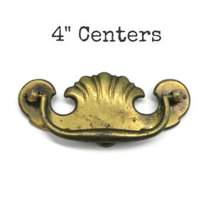 May include: Antique brass drawer pull with a shell design. The pull has a curved handle and decorative backplates. The text "4" Centers" is above the pull. The pull measures 4 inches between the screw holes.