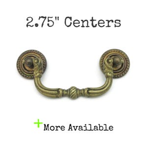 May include: Antique brass drawer pull with a decorative design. The pull has a curved handle and two circular end pieces. The text "2.75" Centers" is above the pull, and "More Available" is below.