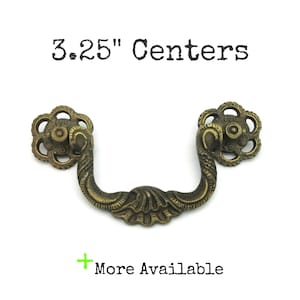 May include: Antique brass drawer pull with a floral design on the ends and a curved handle. The text "3.25" Centers" is above the pull. The text "More Available" is below the pull.