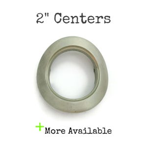 May include: A silver-toned, oval-shaped hardware piece with a 2-inch center. The text "2" Centers" is above the hardware, and "More Available" is below. The hardware has a circular opening in the center.