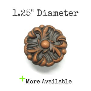 May include: A round copper-toned metal knob featuring a raised floral relief pattern with a central circular detail. The text 1.25" Diameter is printed above the knob, and the text + More Available is printed below the knob on a white background.