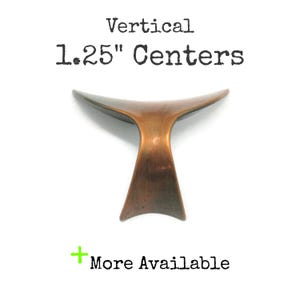 May include: A copper-toned metal cabinet pull with a curved, wing-like shape. The text above the object reads Vertical 1.25" Centers, and the text below reads + More Available. The hardware features a smooth, metallic finish with dark shading.