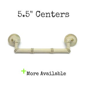 May include: A cream-colored drawer pull with a bamboo-style design and circular fluted mounting plates. The hardware features gold-toned accents and text that reads 5.5" Centers and + More Available.