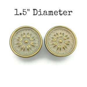 May include: Two antique-style cabinet knobs with a floral design. The knobs are round, with a diameter of 1.5 inches. The knobs have a gold-colored rim and a light-colored center with a raised floral pattern. The text "1.5" Diameter" is above the knobs.