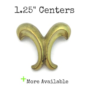 May include: A brass-coloured cabinet pull with a symmetrical, decorative design. The pull measures approximately 3.18 cm across and has curved, ornate details. The text "1.25" Centers" is above the pull, and "More Available" is below.