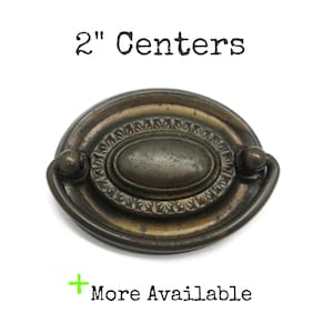May include: An oval-shaped bronze-toned metal drawer pull with a central raised oval plate, a decorative border, and a swinging handle. The text 2" Centers and + More Available is printed in black above and below the hardware piece.