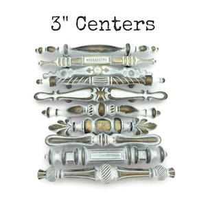 May include: A collection of antique-style cabinet pulls with a distressed white and bronze finish. The pulls are 3 inches (7.6 cm) center-to-center and feature various decorative designs. The text "3" Centers" is displayed at the top.