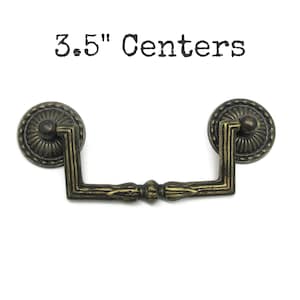May include: An antique brass-toned metal drawer pull with a drop handle design, featuring two circular mounting plates with a ribbed edge and a central bar with decorative detailing. The text 3.5" Centers is printed above the hardware.