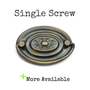 May include: An oval-shaped bronze-toned metal drawer pull featuring a central floral motif surrounded by concentric rings and a beaded border. The text "Single Screw" appears above the hardware, and "+ More Available" is printed below.