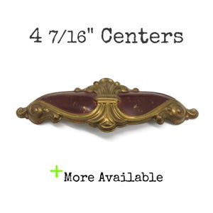 May include: A brass drawer pull with a dark red, distressed inlay featuring ornate scrollwork details. The text 4 7/16" Centers is printed above the hardware, and the text + More Available is printed below the hardware.
