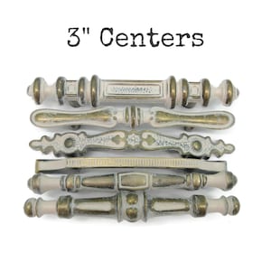 May include: Six decorative cabinet pulls with 3" centers, featuring a mix of off-white and metallic gold finishes. Each handle displays unique ornate detailing, including textured surfaces, cracked paint effects, and varied geometric shapes.