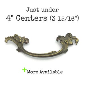 May include: Antique brass-colored drawer pull with ornate, scrolling details. The pull is just under 4 inches (10.16 cm) in length, with the text "Just under 4" Centers (3 15/16")" above the pull. The text "More Available" is below the pull.