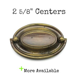May include: Antique brass oval drawer pull with a ribbed centre design. The pull measures 6.67 cm between the centres of the mounting holes. The text "2 5/8" Centers" is above the pull, and "More Available" is below.