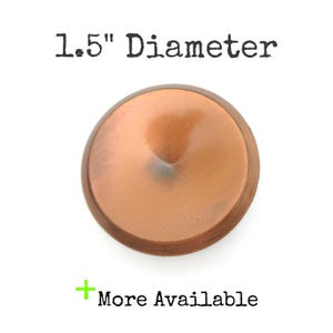 May include: A copper-colored, round knob with a diameter of 1.5 inches. The knob has a slightly domed center and a darker, aged appearance in the center. The text "More Available" is displayed below the knob.