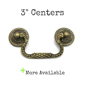 May include: An antique brass-toned metal drawer pull with a swinging handle featuring a diamond-textured center and ornate circular backplates. The text 3" Centers and + More Available is printed above and below the hardware piece.