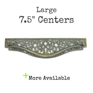 May include: A large antique brass drawer pull featuring an intricate openwork scroll design and a curved handle. The text above reads Large 7.5" Centers, and the text below includes a green plus sign followed by the words More Available.