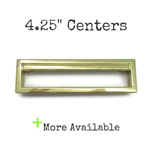 May include: A rectangular gold-toned metal drawer pull with a hollow center. The text 4.25" Centers is printed above the handle, and a green plus sign with the text More Available is printed below the handle.