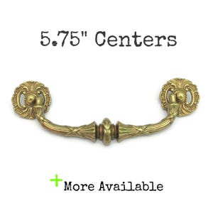 May include: A brass-colored drawer pull with a decorative floral design on each end and a curved handle. The text "5.75" Centers" is above the pull, and "More Available" is below. The pull measures 5.75 inches between the mounting holes.