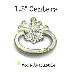 May include: A decorative metal drawer pull featuring a white and brass-toned finish with an ornate floral and scrollwork design. The hardware includes a swinging oval handle and a fixed backplate with the text 1.5" Centers and More Available.