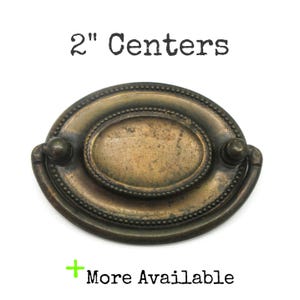 May include: An oval-shaped brass drawer pull with a dark, aged patina and a beaded border detail. The hardware features a central plate with a swinging handle and is labeled with the text 2" Centers and More Available.