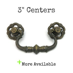 May include: Antique bronze drawer pull with a floral design and a 3-inch (7.6 cm) center-to-center measurement. The pull has a curved handle and decorative end pieces. The text "3" Centers" and "More Available" are visible.