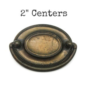 May include: An oval-shaped brass drawer pull with a dark, weathered patina and a beaded border detail. The hardware features a central plate with a swinging handle attached at two points, with the text 2" Centers printed above the object.