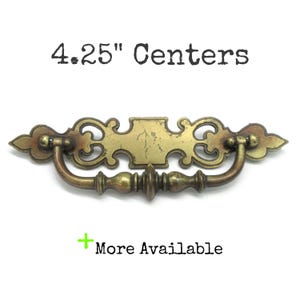 May include: Antique brass drawer pull with a decorative design. The pull has a curved handle and measures 4.25 inches between the centers of the mounting holes. The text "4.25" Centers" is above the pull, and "More Available" is below.