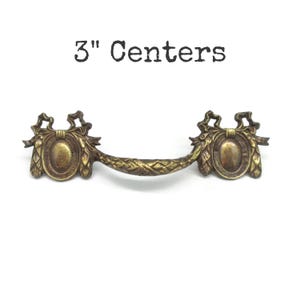 May include: A brass-toned metal drawer pull with a curved handle and two oval-shaped mounting plates featuring ribbon and leaf detailing. The text 3" Centers is printed above the hardware, indicating the distance between the mounting holes.