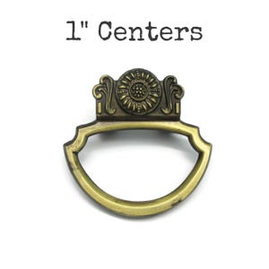 May include: Antique brass drawer pull with a decorative sunflower design and the text "1" Centers". The pull has a curved handle and ornate detailing. The distance between the screw holes is 2.54 cm.