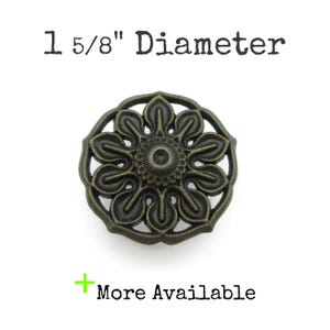 Peut inclure: Bouton de meuble floral de couleur bronze, d'un diamètre de 4,1 cm. Le bouton présente un motif ajouré avec un élément central circulaire et des formes en forme de pétales rayonnant vers l'extérieur. Le texte "More Available" est également visible.