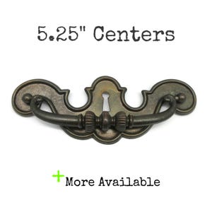 May include: A dark bronze-toned metal drawer pull with a curved handle and a decorative backplate featuring a central keyhole. The text 5.25" Centers and + More Available is printed above and below the hardware piece.