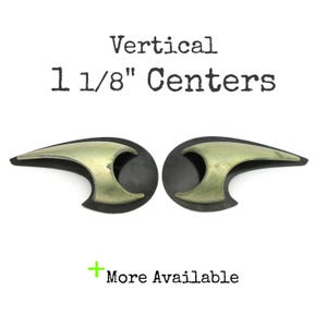 May include: Two decorative cabinet pulls with a brass-colored finish and a black base. The pulls are designed with a unique, curved shape. The text "Vertical 1 1/8" Centers" is printed above the pulls, and "More Available" is printed below.