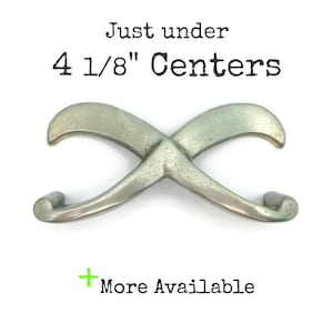 May include: A silver-toned metal cabinet pull with a curved, infinity-symbol design. The text "Just under 4 1/8" Centers" is above the pull, and "More Available" is below. The hardware has a distressed finish.