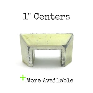 May include: A metallic cabinet pull with a pale yellow finish and a U-shaped design. The text 1" Centers is printed above the pull, and the text + More Available is printed below the pull in black, with a bright green plus sign.