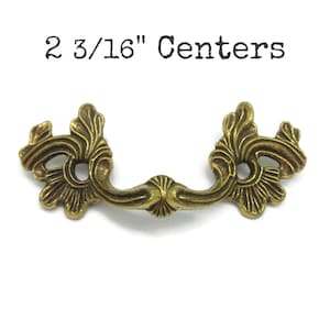 May include: An antique brass-toned metal drawer pull with ornate, symmetrical scrollwork detailing on both sides. The handle features a central grip and is labeled with the text 2 3/16" Centers, indicating the distance between the mounting points.