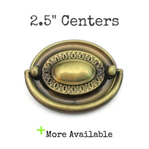 May include: Antique brass-toned drawer pull with an oval center and a decorative border. The pull has a curved handle and measures 2.5 inches between the centers of the mounting holes. The text "2.5" Centers" is above the pull, and "More Available" is below.