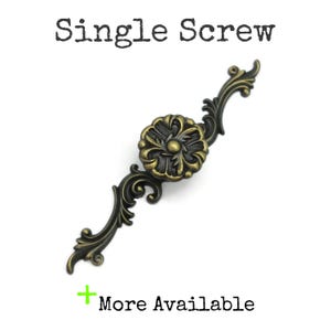 May include: An antique brass-colored metal drawer pull featuring a central circular floral motif with ornate, scrolling leaf-like extensions on both sides. The text "Single Screw" appears above the hardware, and "+ More Available" is below.