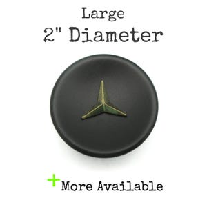 May include: A black, round object with a bronze-colored starburst design in the center. The text "Large 2" Diameter" is above the object, and "More Available" is below it.