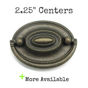 May include: An oval-shaped antique brass drawer pull with a beaded border and a central raised oval detail. The hardware features a swinging handle and is labeled with the text 2.25" Centers and More Available.