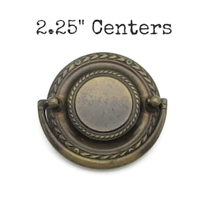May include: Antique brass-toned drawer pull with a circular design. The pull features a central disc surrounded by a rope-like texture and a decorative outer ring. The text "2.25" Centers" is printed above the pull.