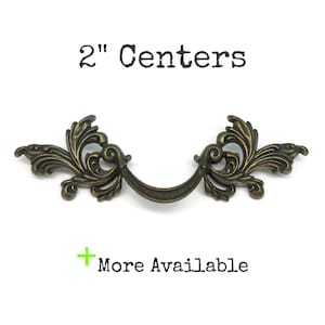 May include: A bronze-toned metal drawer pull with ornate, scrolling leaf-like details on both sides and a curved central handle. The text 2" Centers is printed above the hardware, and a green plus sign with the text More Available is below.