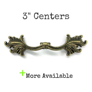 May include: An antique brass-toned metal drawer pull featuring ornate, leaf-like decorative ends and a central shell motif. The text above reads 3" Centers and the text below includes a green plus sign followed by the words More Available.