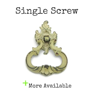May include: An antique-style drawer pull with a distressed, off-white finish. The pull features an ornate design with a loop handle and decorative details. The text "Single Screw" is above the pull, and "More Available" is below.